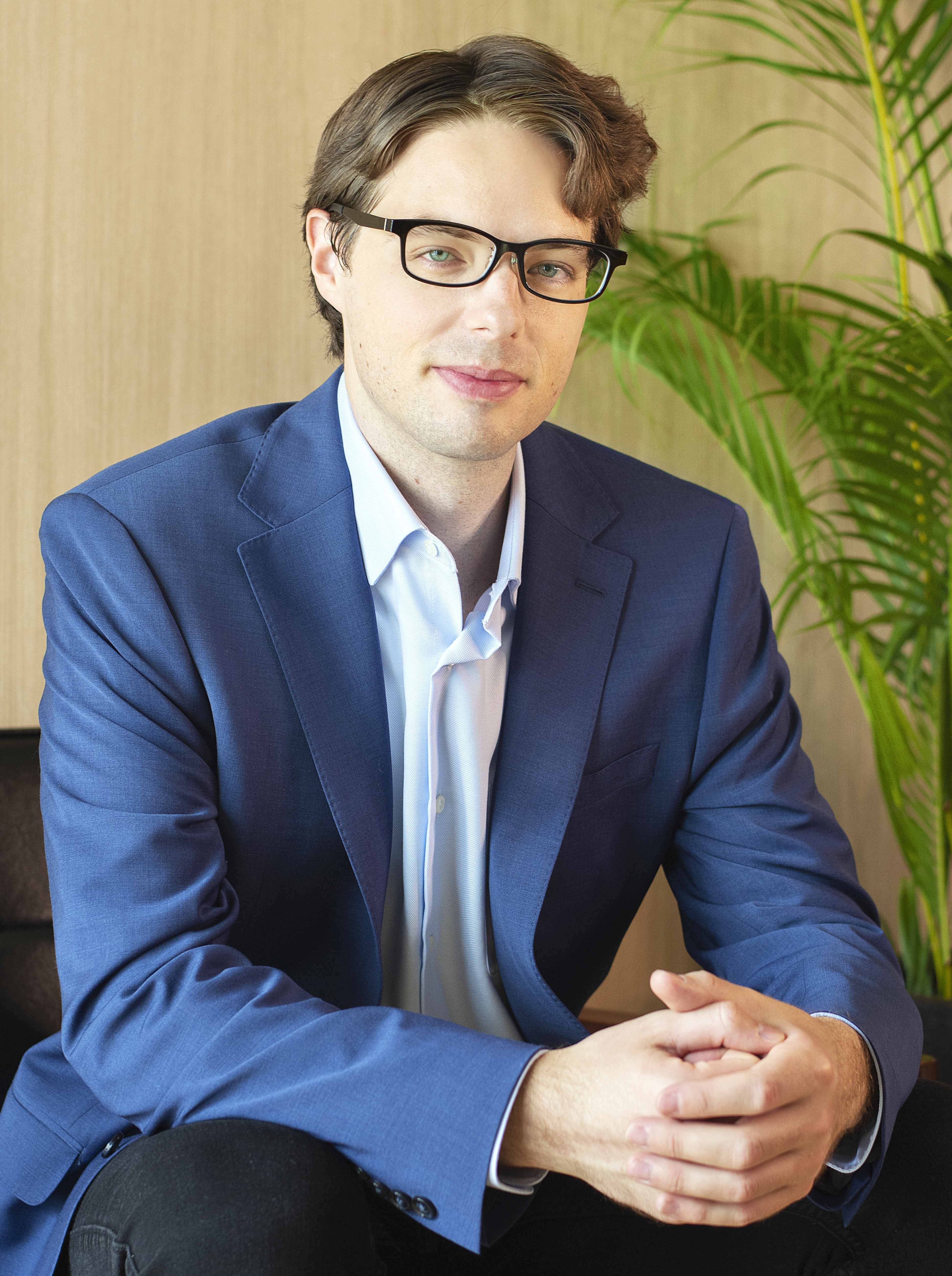 Photo of William Franklin sitting on a sofa wearing a blue suit in an office smiling